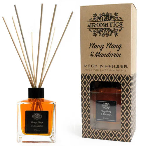 A "Ylang Ylang & Mandarin" reed diffuser bottle and its brown cardboard box displayed side-by-side on a white background. The bottle holds an orange-colored liquid, and the box has a window revealing the bottle inside.