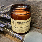 Brown glass aromatherapy candle jar labeled "Positive Vibes," placed on rustic wooden surface with lavender flowers beside it.