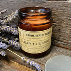 Aromatherapy candle in a brown glass jar labeled "Clear Thinking," placed on rustic wood with lavender flowers beside it.