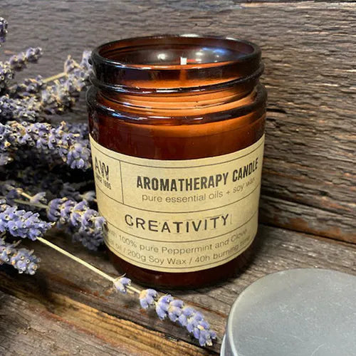 Amber glass jar candle labeled “Aromatherapy Candle – Creativity” placed on rustic wooden surface with lavender sprigs and lid nearby; text highlights peppermint and clove essential oils.