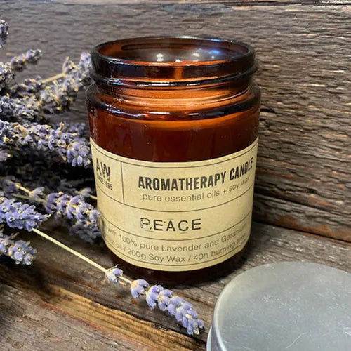 Brown glass aromatherapy candle in rustic setting with lavender sprigs, labeled “Peace” and made with pure lavender and geranium essential oils, soy wax, 40h burning time.
