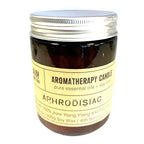 Aromatherapy candle in amber glass jar with silver lid and label reading “Aromatherapy Candle – Aphrodisiac,” made with pure Ylang Ylang and Patchouli essential oils.