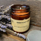 Open amber glass aromatherapy candle labeled “Aphrodisiac” placed on rustic wooden surface with sprigs of lavender beside it and the silver lid resting nearby.