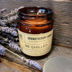 Open "Be Chilled" aromatherapy candle in brown glass jar, placed on rustic wood with lavender sprigs beside it.