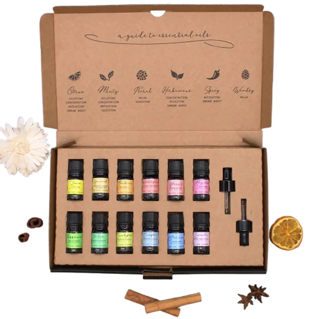 Essential oils gift set displayed inside a cardboard box, containing 12 small brown glass bottles with colorful labels, two droppers, and decorative dried flowers and spices around the box.