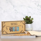 A product shot of a brown cardboard gift box for an aromatherapy eye pillow with a blue and black spotted design. The box is placed on a marbled surface next to a plant, with wheat seeds spilling from the inner pouch, highlighting the natural elements of this home fragrance and aromatherapy product.