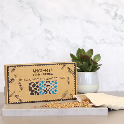 A product shot of a brown cardboard gift box for an aromatherapy eye pillow with a blue and black spotted design. The box is placed on a marbled surface next to a plant, with wheat seeds spilling from the inner pouch, highlighting the natural elements of this home fragrance and aromatherapy product.