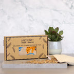 A modern product shot showing a gift box for an aromatherapy eye pillow with a "Lazy Sloth" pattern. The box is placed on a marble surface next to a plant, with wheat seeds spilling from the inner pouch, emphasizing the natural elements of this home fragrance and aromatherapy product.