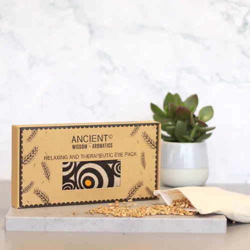 A modern product shot showing a gift box for an aromatherapy eye pillow with a black, white, and orange spiral "Illusion" pattern. The box is placed on a marble surface next to a plant, with wheat seeds spilling from the inner pouch, emphasizing the natural elements of this home fragrance and aromatherapy product.