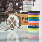 Silver essential oil diffuser necklace with four-leaf clover heart design, displayed with colorful felt pads and essential oil bottle in background.