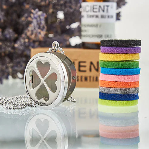 Silver essential oil diffuser necklace with four-leaf clover heart design, displayed with colorful felt pads and essential oil bottle in background.