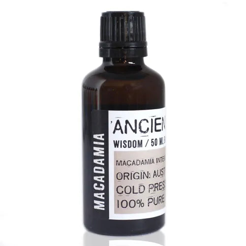 Amber glass bottle labeled “Ancient Wisdom / 50 ml Base Oil.” The label specifies “Macadamia Integrifolia” (Macadamia oil), origin Australia, cold pressed, and 100% pure oil. The main label area is light beige with bold black text, while the vertical side strip reads “MACADAMIA” in white on a black background. The bottle has a black ribbed screw cap, presenting a clean apothecary-style design.
