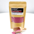 Brown kraft pouch of Ancient Wisdom Aromatherapy Bath Potion labeled “Passion Potion” with Ylang Ylang, Lemon & Orange essential oils, showing the front label and transparent window filled with pink bath salts, with a small wooden scoop spilling pink salts in the foreground.