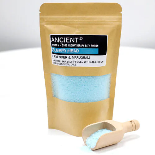 Kraft paper pouch of "Sleepy Head" bath salts with blue granules and a wooden scoop spilling some salt on a white background.