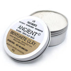 Open round metal tin of Ancient Wisdom Earth Clay Mask labeled "BENTONITE CLAY." The lid, placed at an angle, shows the label highlighting antibacterial and anti-inflammatory properties, made in the United Kingdom, 80g. The container reveals fine, light-colored bentonite clay powder inside.