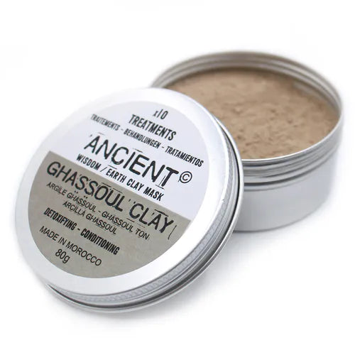 Open silver metal tin containing Ancient Wisdom Ghassoul Clay mask powder. The label on the lid reads "Ancient Wisdom / Earth Clay Mask – Ghassoul Clay," with descriptions in multiple languages and benefits "Detoxifying – Conditioning." The clay inside the tin appears light brown and finely ground. The product is made in Morocco, 80g, and is marketed for skincare treatments.