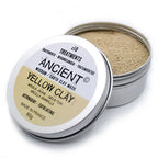 Open silver tin container of “Ancient Wisdom Earth Clay Mask – Yellow Clay.” The lid rests slightly tilted, showing the label that reads “x10 Treatments,” “Yellow Clay” in bold, with translations in French, German, and Spanish. The text highlights benefits “Astringent – Exfoliating, Made in France, 80g.” The container is filled with light yellow-beige clay powder with a fine, grainy texture visible on the surface.
