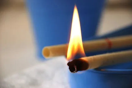 Close-up of two ear candles, one burning with a bright flame while placed over a bowl, with blurred background in blue tones.