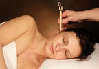 A woman lying on her side with eyes closed during an ear candling session, while a practitioner holds a lit ear candle gently in her ear.