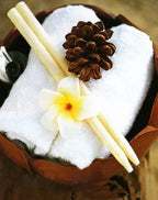 Decorative spa setting with two ear candles placed on a rolled white towel inside a wooden bowl, accented with a pine cone and a white tropical flower.
