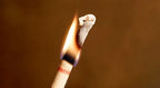 Close-up of a hollow ear candle being lit at the tip, with a visible flame and burnt edges against a plain background.