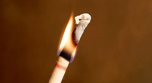 Close-up of a hollow ear candle being lit at the tip, with a visible flame and burnt edges against a plain background.