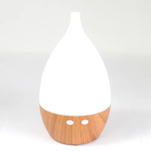 A product shot of a teardrop-shaped essential oil diffuser with a tall white top and a rounded light wood grain base. It is on a white background.