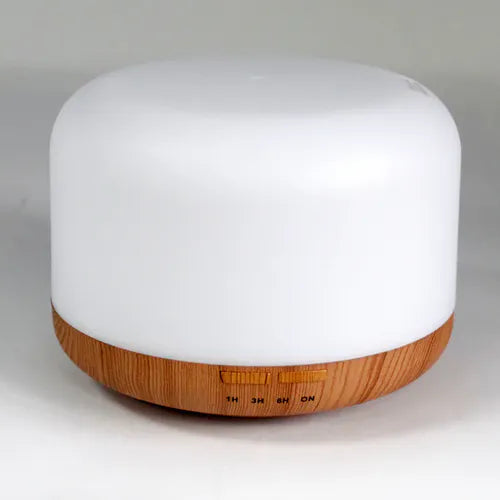 A product shot of a large, cylindrical essential oil diffuser with an opaque white top that lights up and a light wood grain base. The base has buttons for the 1-hour, 3-hour, and 6-hour timers, as well as an ON button.