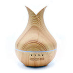 A product shot of a tall essential oil diffuser shaped like a flower bud, with a light wood grain base and a white top. It is on a white background.