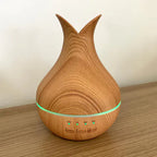 A close-up, front-facing shot of an essential oil diffuser on a wooden surface. The diffuser has a light wood grain base and a tall, pointed top. A green light is visible in the middle band, and timer and power buttons are visible at the bottom.