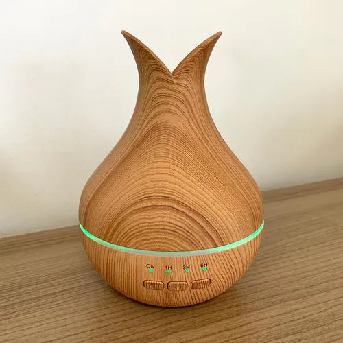 A close-up, front-facing shot of an essential oil diffuser on a wooden surface. The diffuser has a light wood grain base and a tall, pointed top. A green light is visible in the middle band, and timer and power buttons are visible at the bottom.