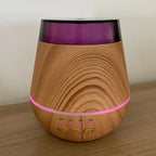 Wood grain aroma diffuser placed on a wooden surface, illuminated with a pink LED ring light and active timer indicators for ON, 1H, 3H, and 6H beneath three control buttons.
