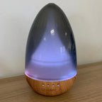 Aroma diffuser with a glowing LED light in shades of purple, shaped like a teardrop with a wooden base and control buttons (ON, 1H, 3H, 6H) visible at the bottom.