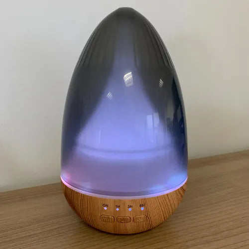 Aroma diffuser with a glowing LED light in shades of purple, shaped like a teardrop with a wooden base and control buttons (ON, 1H, 3H, 6H) visible at the bottom.
