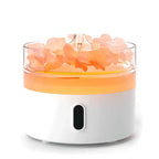 Round white aroma diffuser with transparent top filled with pink crystal-like stones, illuminated with orange light against a plain background.