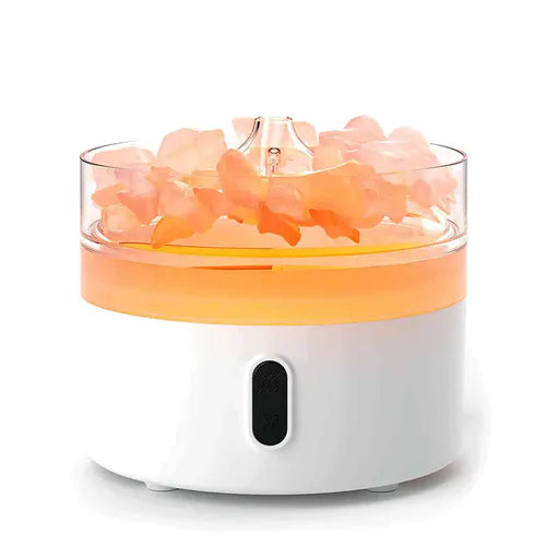 Round white aroma diffuser with transparent top filled with pink crystal-like stones, illuminated with orange light against a plain background.