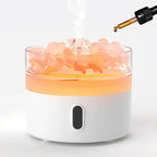 Round white aroma diffuser with transparent top filled with pink crystal-like stones, emitting steam while a dropper adds essential oil from above.