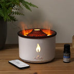 White cylindrical aroma diffuser with glowing orange light and flame effect from the top, placed on a wooden surface next to a remote control and a lavender essential oil bottle.