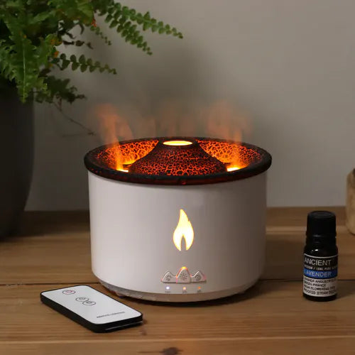White cylindrical aroma diffuser with glowing orange light and flame effect from the top, placed on a wooden surface next to a remote control and a lavender essential oil bottle.