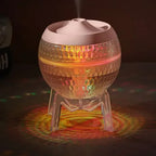 Spherical glass aroma diffuser with a pink lid, lit up with warm yellow, red, and green LED lights, placed on a clear tripod stand.