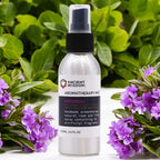A 100ml aluminum spray bottle of "Ancient Wisdom Aromatherapy Mist Patchouli" with a black spray nozzle and white label, positioned outdoors among fresh green leaves and vibrant purple flowers, creating a natural and aromatic setting.