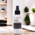 A 100ml silver aluminum spray bottle of "Ancient Wisdom Aromatherapy Mist - Ylang Ylang & Mandarin," placed on a white surface with a blurred cozy indoor setting in the background, including plants and shelves, emphasizing a natural and relaxing atmosphere.