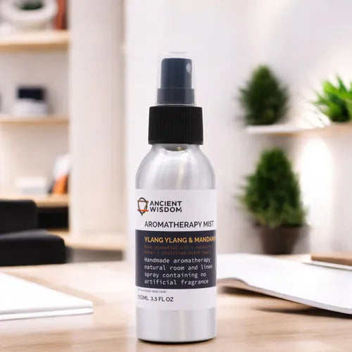 A 100ml silver aluminum spray bottle of "Ancient Wisdom Aromatherapy Mist - Ylang Ylang & Mandarin," placed on a white surface with a blurred cozy indoor setting in the background, including plants and shelves, emphasizing a natural and relaxing atmosphere.