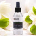 Aluminum spray bottle labeled “Ancient Wisdom Aromatherapy Mist – May Chang & Bergamot.” The 100ml (3.5 fl oz) bottle has a white label with black and green text, black spray cap, and is displayed among soft white flowers and green leaves, creating a fresh and natural atmosphere.