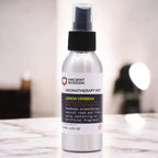 A 100ml aluminum spray bottle of Ancient Wisdom Aromatherapy Mist in the Lemon Verbena scent, shown on a marble surface with a blurred indoor background. The bottle has a black spray cap and a white label with black and yellow text. The label indicates it is a handmade natural room and linen spray made with pure essential oil, Yorkshire water, and distilled witch hazel, with no artificial fragrance.