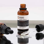 "Amber glass dropper bottle of Ancient Wisdom Facial Serum with Vitamin C, labeled 'Bright & Healthy.' The label highlights Aloe Vera and Witch Hazel as key ingredients to stimulate collagen production and promote radiant skin. The 30ml bottle is made in England and designed for all skin types. The bottle is photographed upright with the dropper removed, lying on the reflective surface with serum droplets visible."