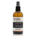 Brown glass spray bottle labeled "Ancient Wisdom Facial Toner Mist Hydrating Natural," featuring witch hazel extract. The white and black label indicates it is hydrating and soothing for all skin types. Bottle shown upright with black spray cap.