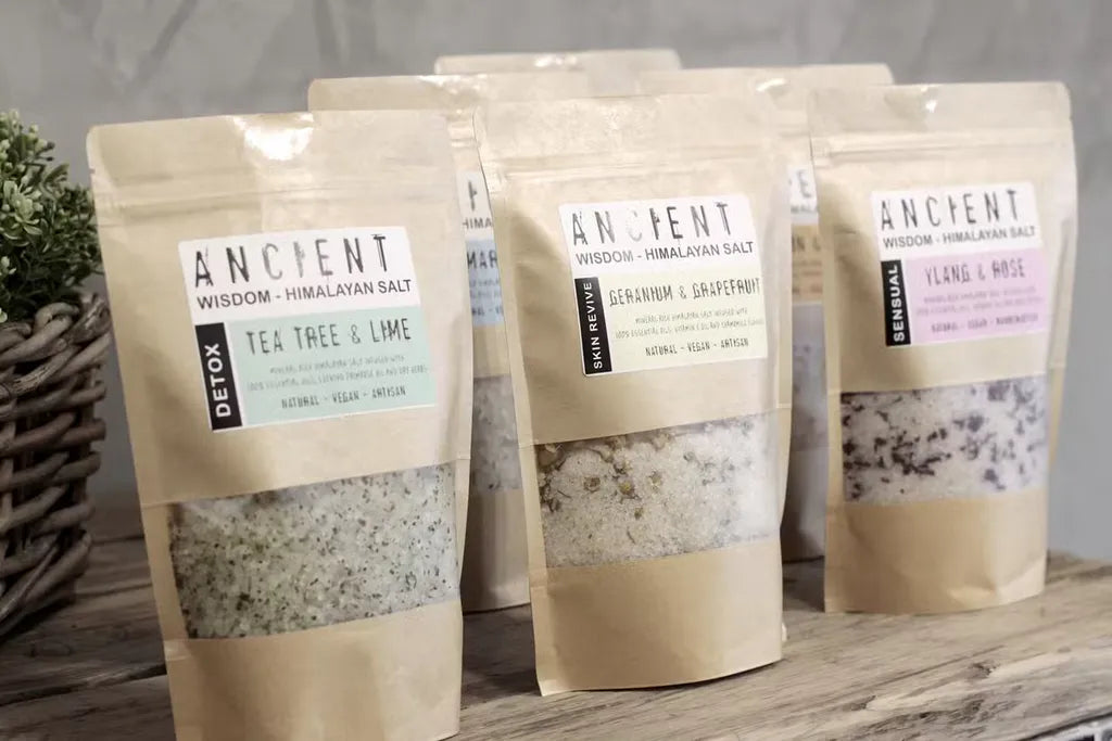 A selection of Ancient Wisdom Himalayan bath salts in kraft paper pouches, displayed on a wooden surface. Labels indicate different blends such as Tea Tree & Lime (Detox), Geranium & Grapefruit (Skin Revive), and Ylang & Rose (Sensual). The salts are visible through the transparent windows of the packaging.