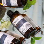 Group of amber glass bottles of Ancient Wisdom Floral Waters lying on a reflective surface, including Geranium, Lavender, and other variants, decorated with illustrated floral motifs.