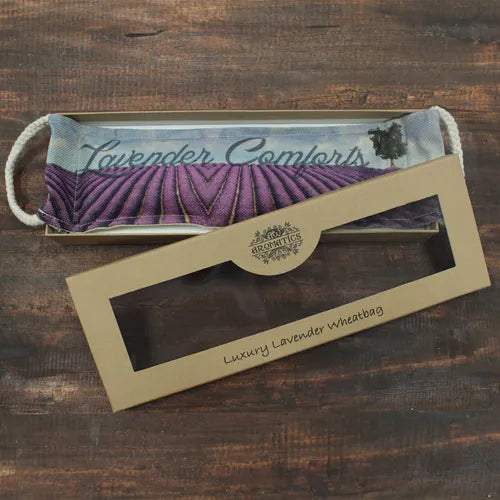 An unboxed lavender wheat bag placed on a wooden surface next to its open cardboard packaging. The bag displays a printed lavender field with the words “Lavender Comforts,” designed for warmth and relaxation.
